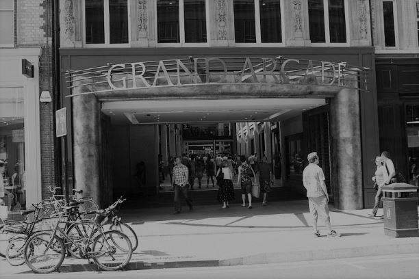 The frontage of the Grand Arcade in Cambridge. Large letters spell out Grand Arcade with shoppers milling about below.